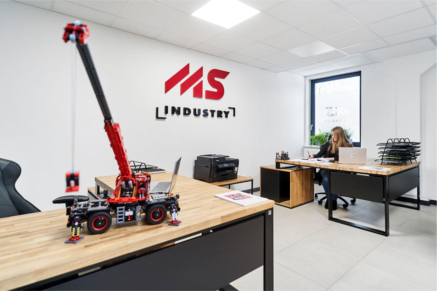 Biuro MS Industry z logo na ścianie, stanowiska pracy oraz model żurawia na biurku. 